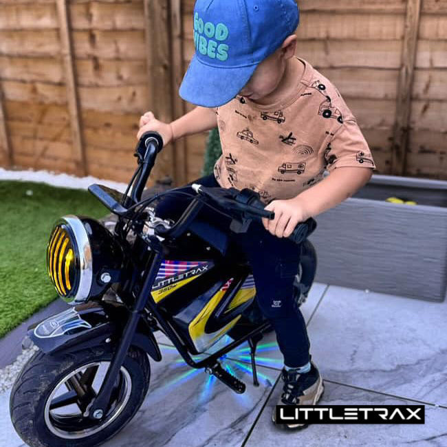 Child on a toy motorcycle with 'LITTLETRAX' branding, wearing a blue cap and patterned shirt.