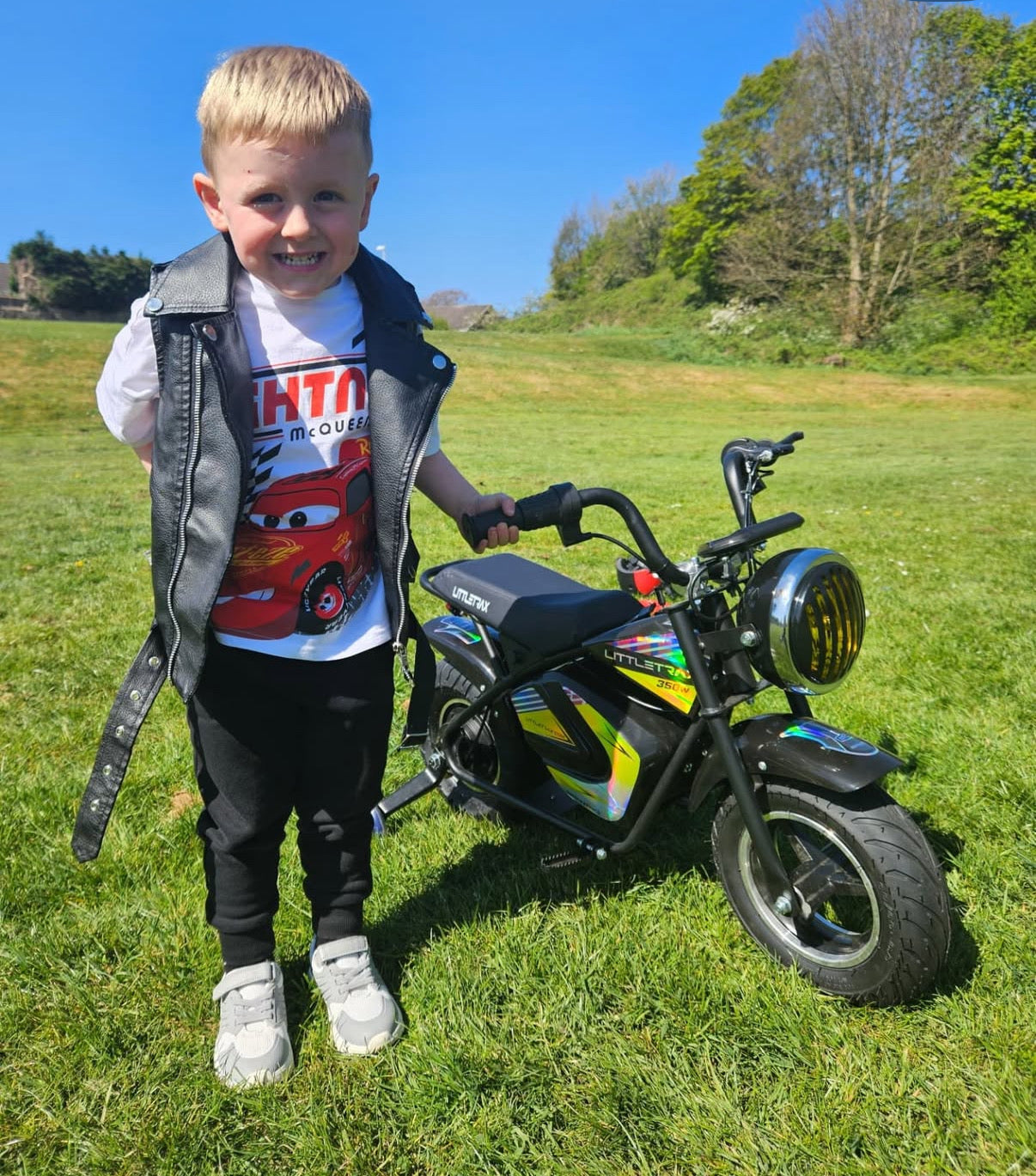Littletrax 350W Kids Electric Bike Child standing next to a small motorcycle on a grassy field with trees in the background