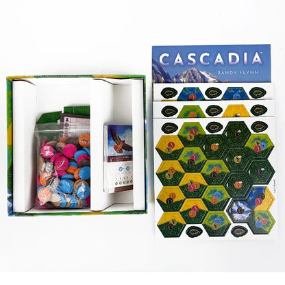 Board game components including hexagonal tiles and a box with 'Cascadia' branding.