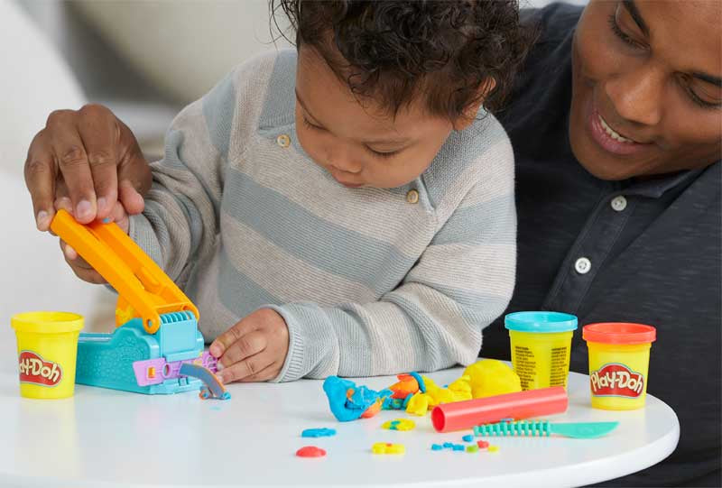 Child playing with Play-Doh toys at a table with an adult next to them.