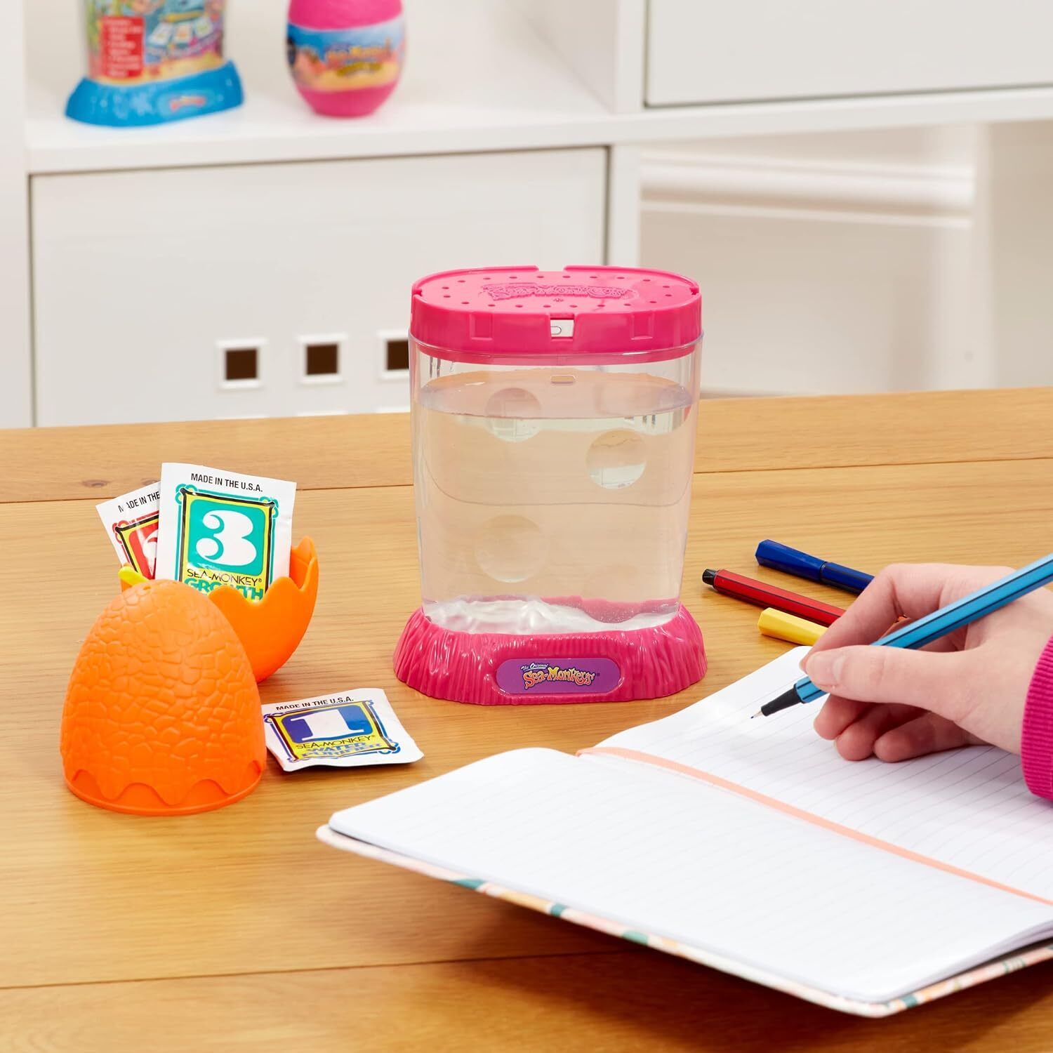 The Original Sea Monkeys - Ocean Zoo Grow Your Own Pets Science Kit 6+ years Person writing in a notebook with a pink container and orange object on a wooden surface
