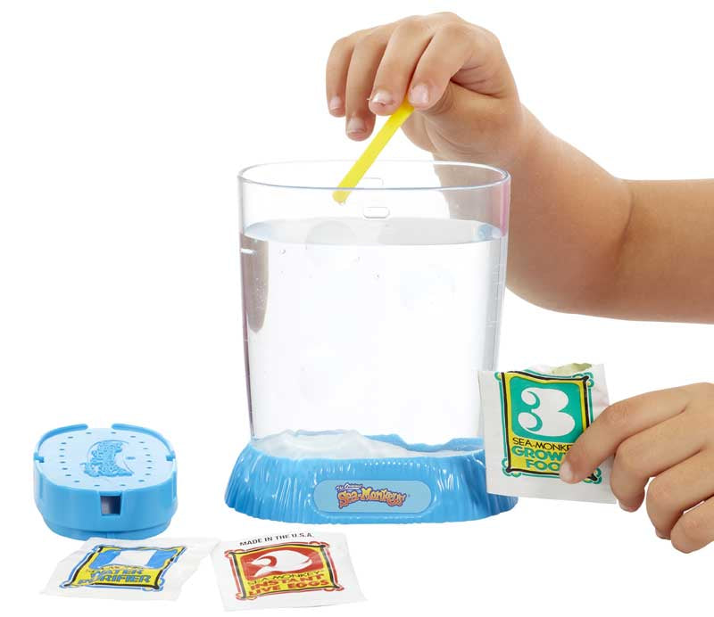 The Original Sea Monkeys - Ocean Zoo Grow Your Own Pets Science Kit 6+ years Child's hand stirring a glass of water with a yellow straw, next to a blue device and packets on a white background.